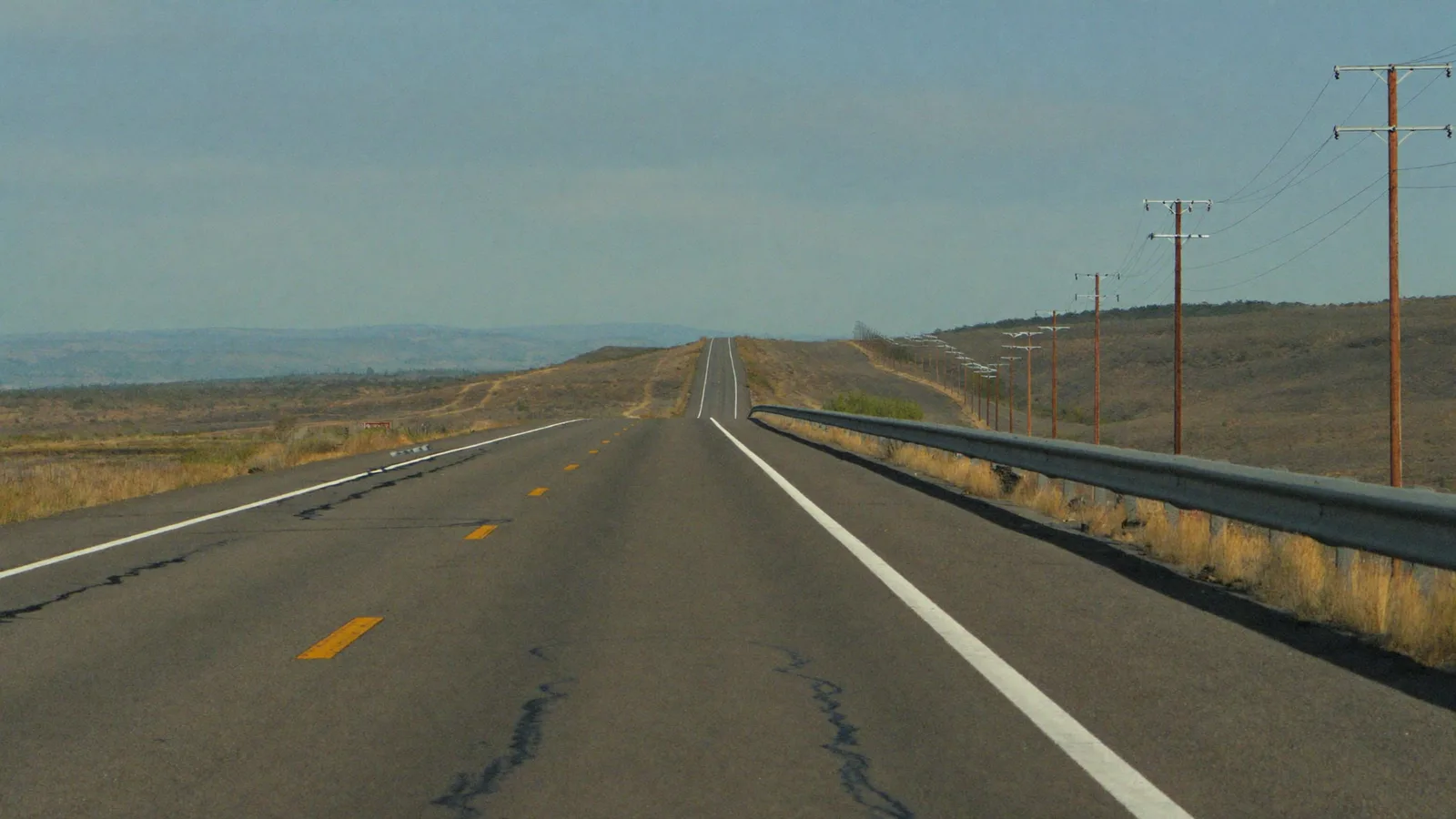 An empty two-lane US rural highway running toward low hills, with distribution poles alongside