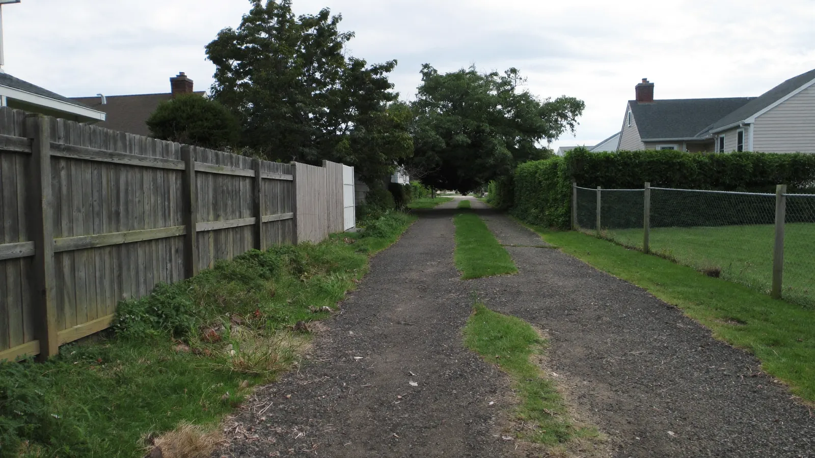 A long shared gravel and asphalt driveway running between two US suburban properties, a wood privacy fence on one side and a hedge on the other