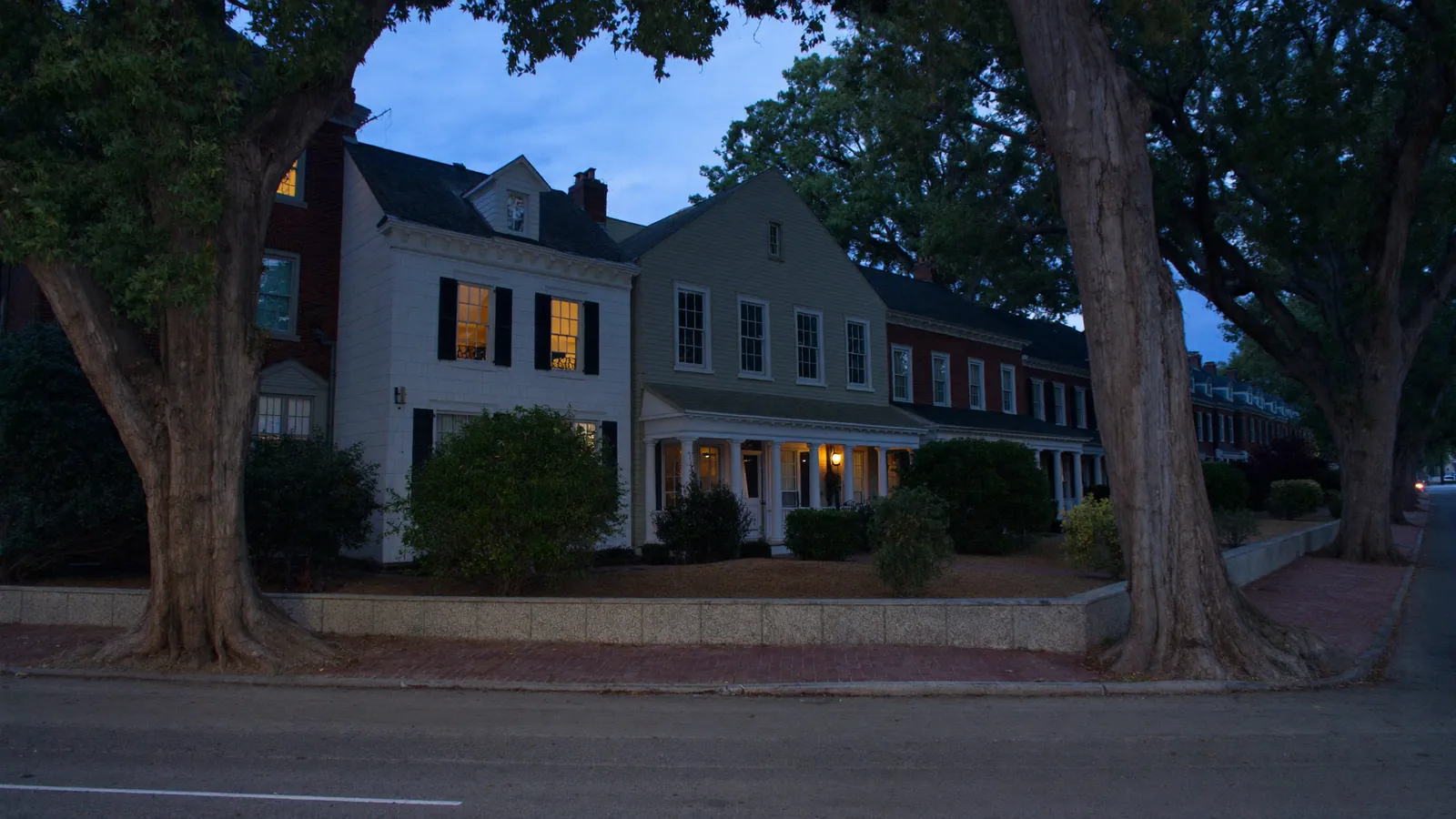 A historic US residential street at blue hour, colonial-revival houses with columned porches behind mature street trees and a brick sidewalk
